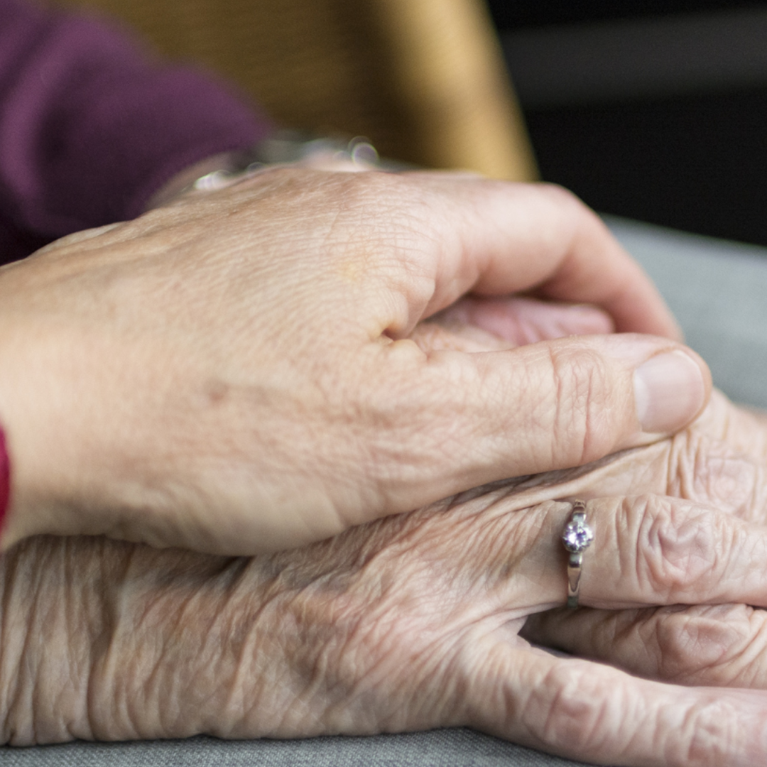 a close up shot of an elderly couple holding hands in a comforting gesture. the hand underneath is wearing a diamond wedding band 